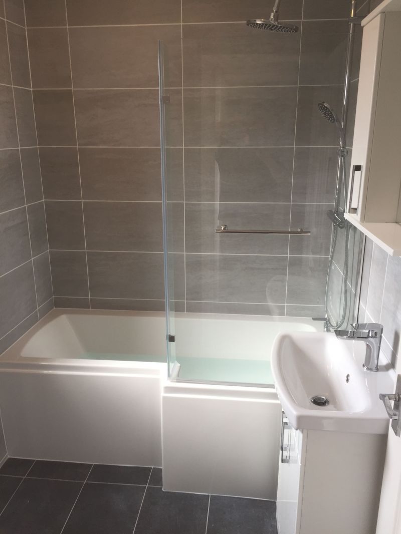 A modern bathroom suite with shower door and over bath shower with Grey wall and floor tiling. : Swipe To View More Images