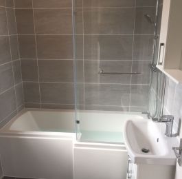 A modern bathroom suite with shower door and over bath shower with Grey wall and floor tiling. : Click Here To View Larger Image